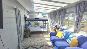 Una habitación iluminada por el sol con una litera, un sofá azul con cojines, una alfombra estampada y una planta en maceta.