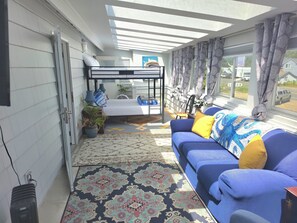 A sunlit room with a bunk bed, a blue sofa with cushions, a patterned rug, and a potted plant.