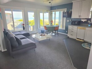 Sala de estar com sofá cinzento, mesinha de centro e área de jantar com mesa e cadeiras. Cozinha com armários brancos e lava-louças. Grandes janelas oferecem vista para o oceano.