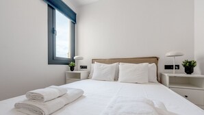 A neatly made bed with white linens and two rolled towels, a wooden headboard, a window with blinds, bedside tables with lamps and small plants, and a white wall. - GA1 (Barcelona)