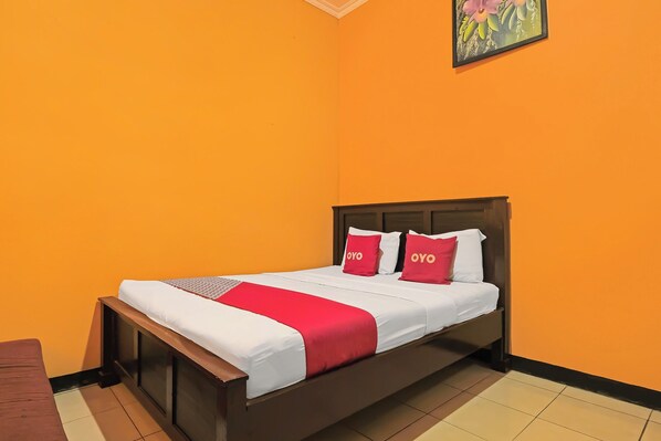 Una cama individual con estructura de madera, ropa de cama blanca y detalles en rojo, en una habitación con paredes anaranjadas y suelo de baldosas.