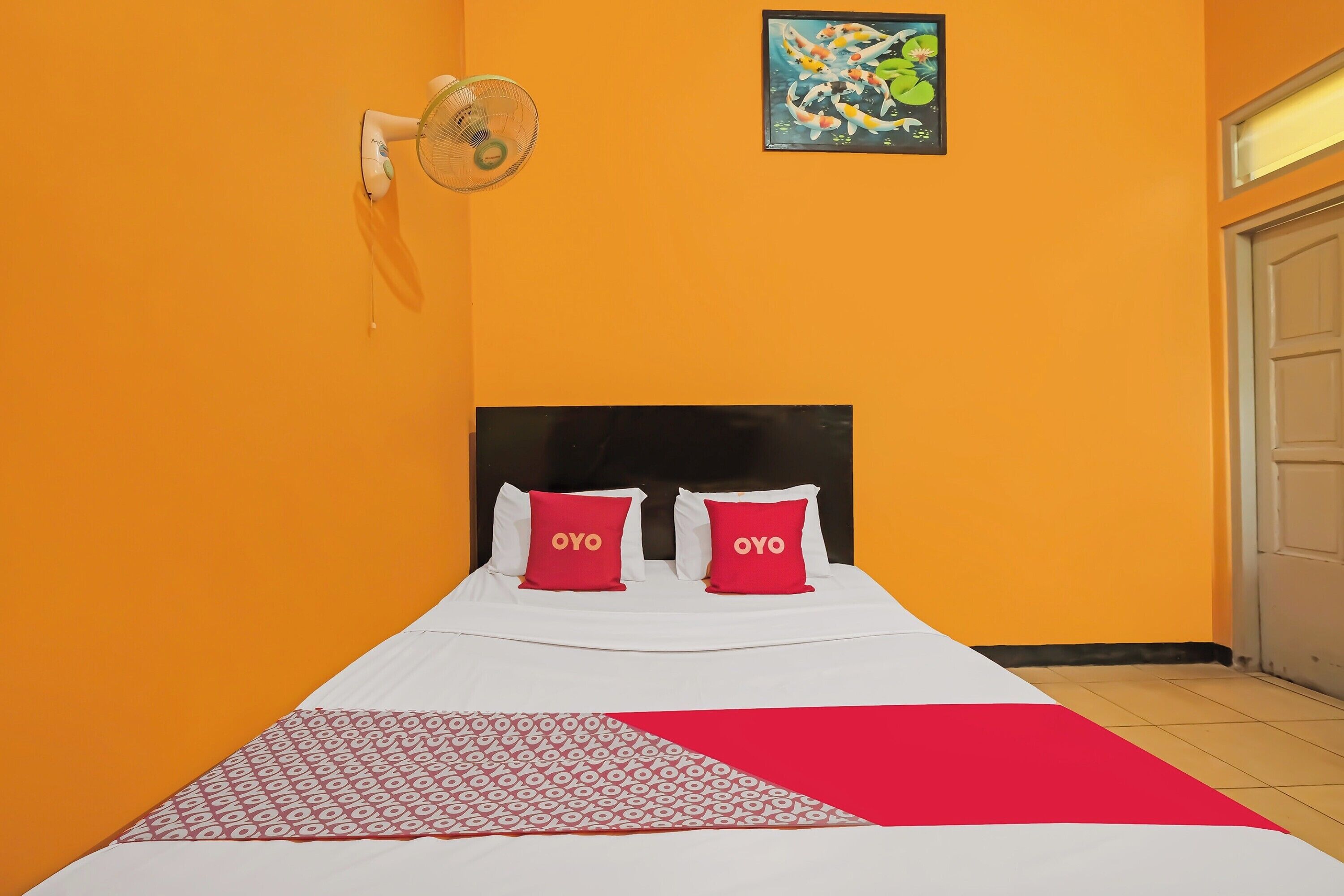 Habitación de hotel con pared naranja, cama con ropa de cama blanca y almohadas rojas, cabecero negro, ventilador fijado a la pared y un cuadro enmarcado sobre la cama.