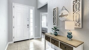 Un pasillo con una puerta blanca, una consola decorativa con adornos y una pared texturizada con un cartel de bienvenida.