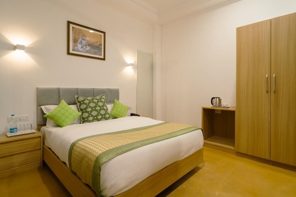 Una cama bien hecha con una colcha verde y blanca de estampado, un cabecero gris y dos lámparas de noche. A la derecha, un armario de madera y una mesita con un hervidor y una taza.