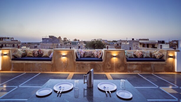 Un espacio de comedor en la azotea con vistas a la ciudad, una zona de estar con cojines y una mesa dispuesta con platos y cubiertos.