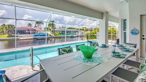 Area pranzo a bordo piscina con vista su un porto turistico, un tavolo bianco con sottopiatti verdi e una ventola a soffitto.