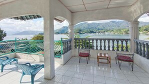Varanda coberta com vista para um lago e montanhas, com várias cadeiras e uma mesa.