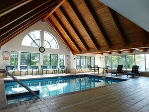 An indoor swimming pool with wooden beams, large windows, and chairs around the pool area.