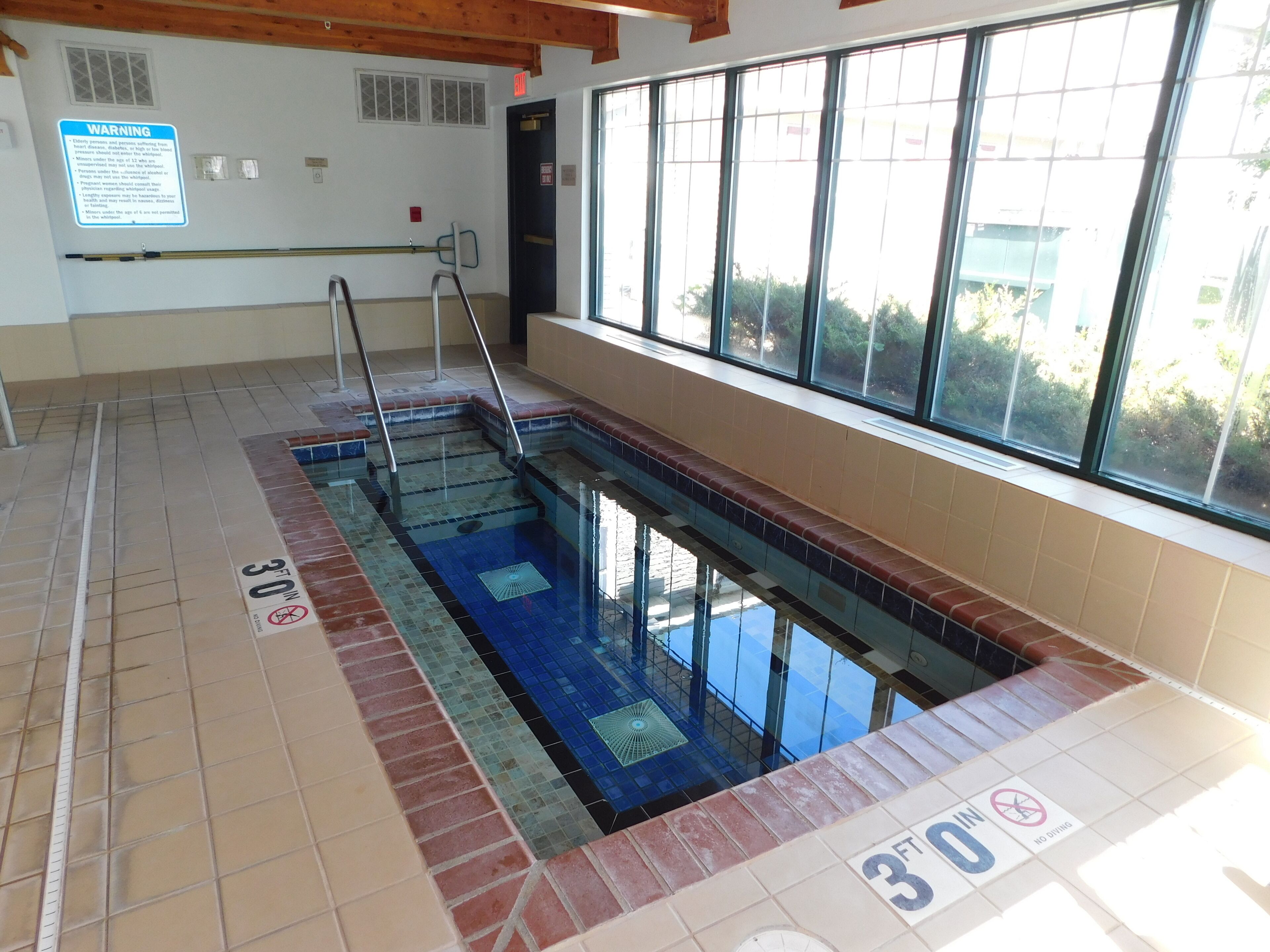 Une piscine intérieure dont la profondeur maximale est indiquée à 3 pieds, avec un panneau d’avertissement sur le mur et une fenêtre donnant sur un espace verdoyant à l’extérieur.