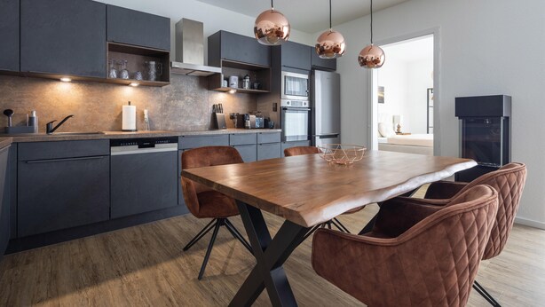 A modern kitchen with dark cabinets, a wooden dining table, and copper pendant lights. - Park - Apartment 12 im Marinapark Scharmützelsee, Sauna, Whirlpool, E - Kamin, 4 Pers., 80 m² (Wendisch Rietz Siedlung)