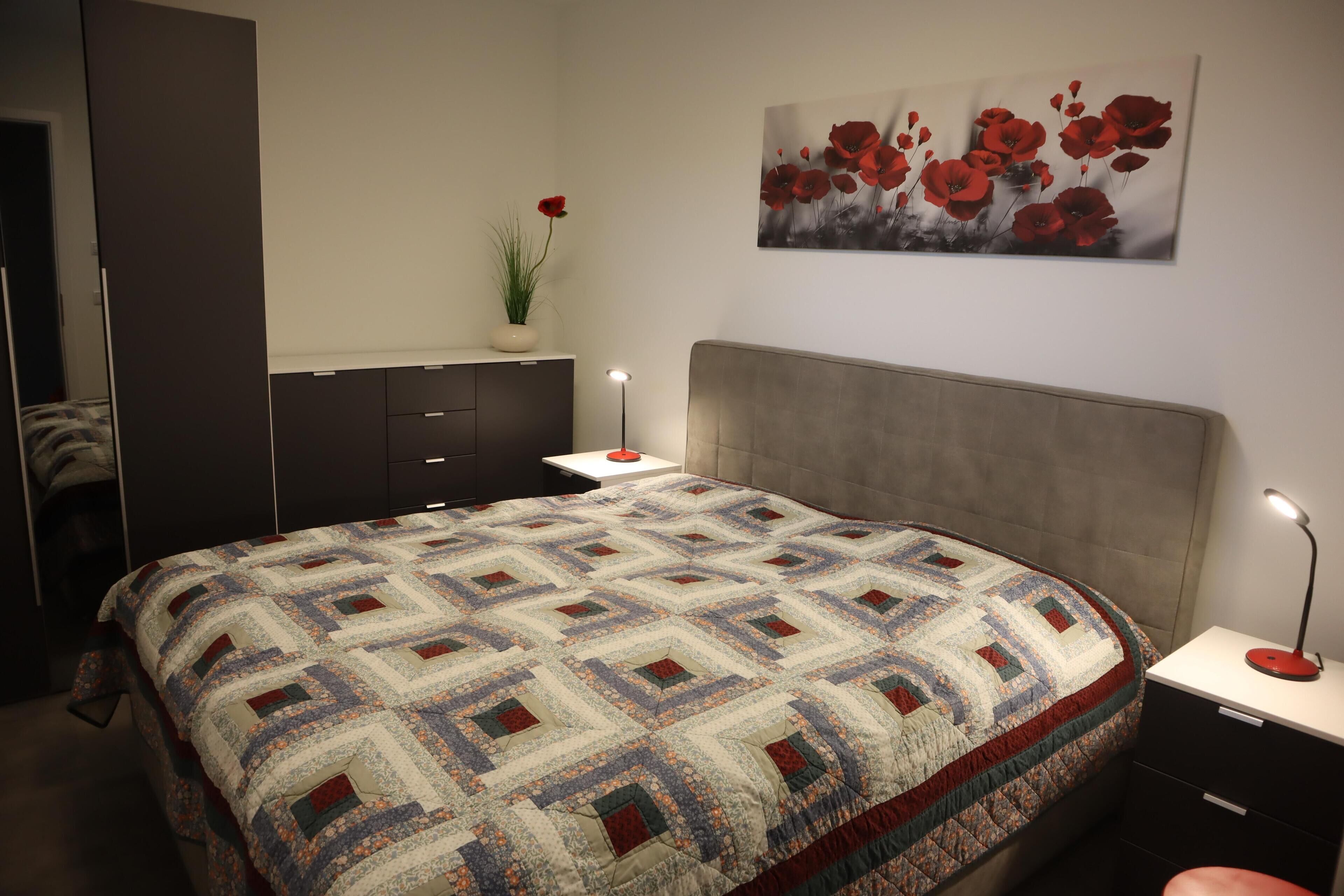 Ein Schlafzimmer mit einem großen Bett, einem Nachttisch, einer Kommode, einem Gemälde von roten Blumen und einer Topfpflanze.