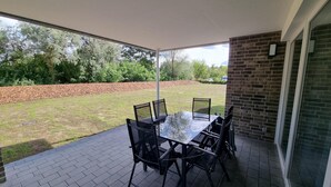An outdoor patio with a table and chairs, a brick floor, and a view of a grassy area with trees and a brick wall. - Park - Apartment 01 im Marinapark Scharmützelsee, Sauna, Whirlpool, 6 Pers., 98 m² (Wendisch Rietz Siedlung)
