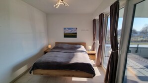 Une chambre avec un lit en bois, deux tables de chevet avec des lampes, une vue sur un parc grâce à de grandes fenêtres, et un tableau accroché au mur.