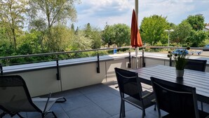Une terrasse extérieure avec une table et des chaises, un pot de fleurs et une vue sur un parking et des espaces verts.