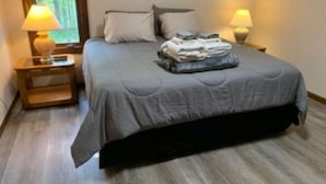 A neatly made bed with a grey bedspread and pillows, two bedside lamps, and a window with a view of trees.