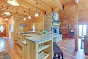 A wooden cabin interior with a kitchen island, stone fireplace, and wooden ceiling beams.