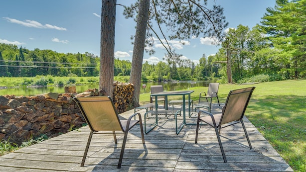 En træterrasse med udendørs møbler, en stenvæg og udsigt til en sø omgivet af træer.
