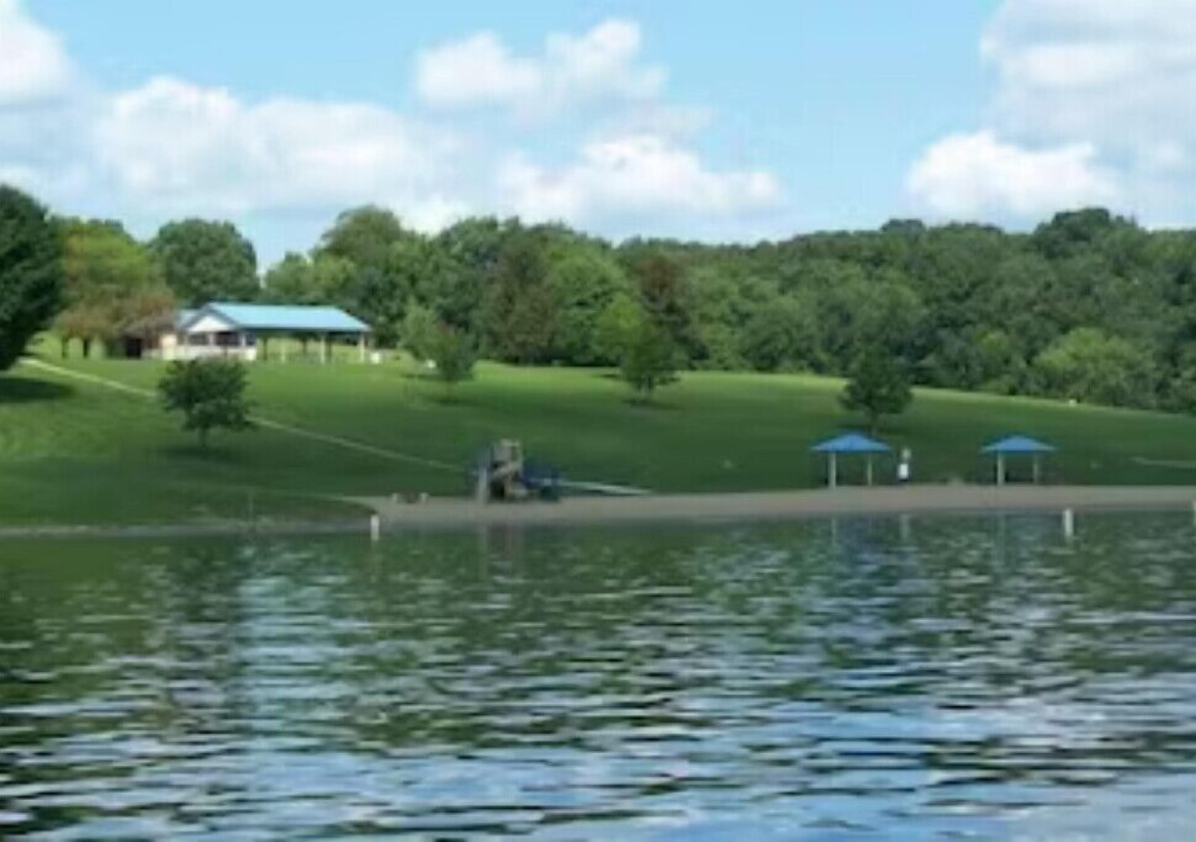 Blick auf einen See mit einem Pavillon, einer überdachten Fläche und einem Gebäude im Hintergrund.