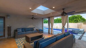 Ein modernes Wohnzimmer mit einer großen Couch, einem Couchtisch und einem Deckenventilator. Eine Glastür führt zu einem Poolbereich.