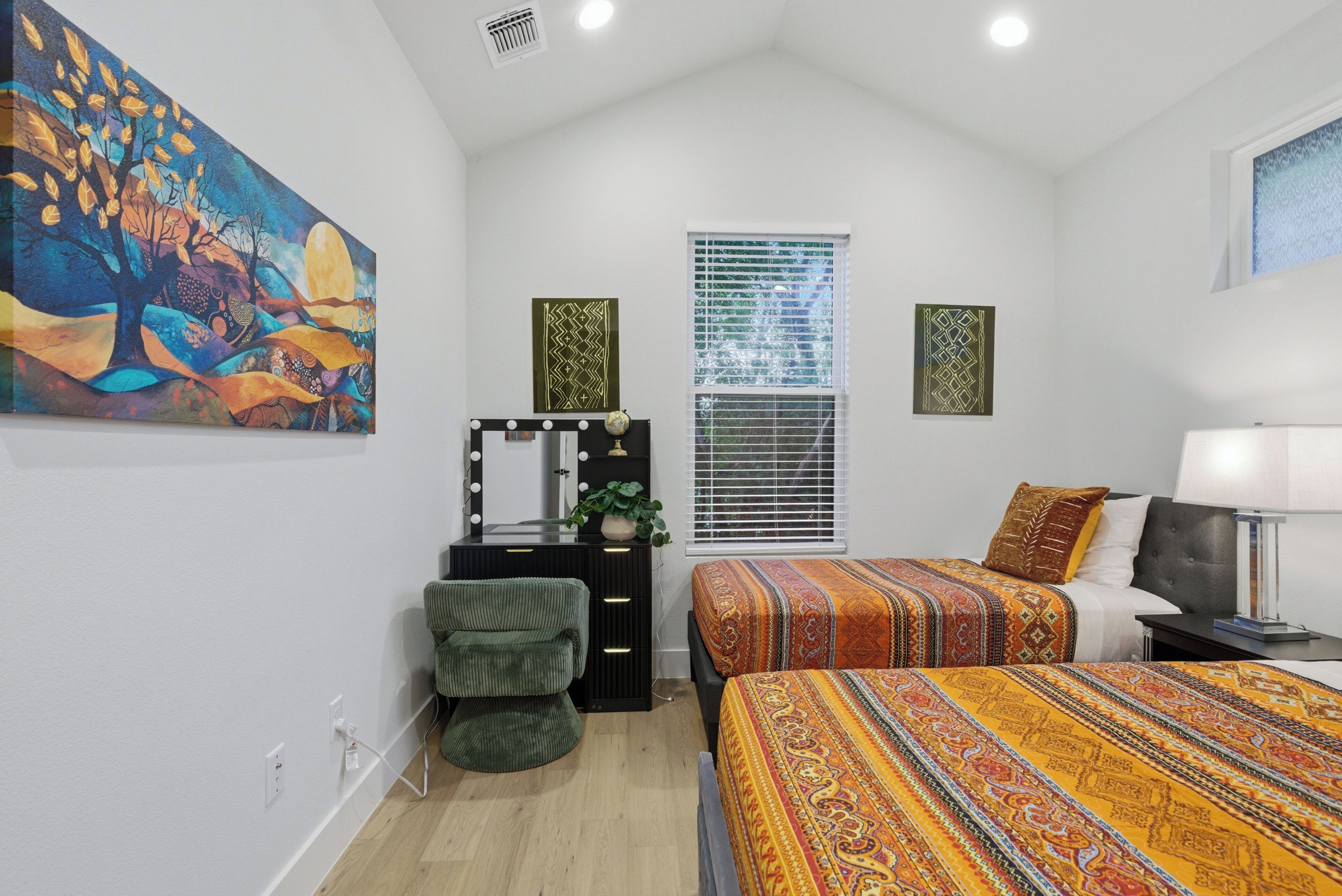 A bedroom with a bed, a dresser, a vanity with mirrors, a window with blinds, and a painting on the wall.