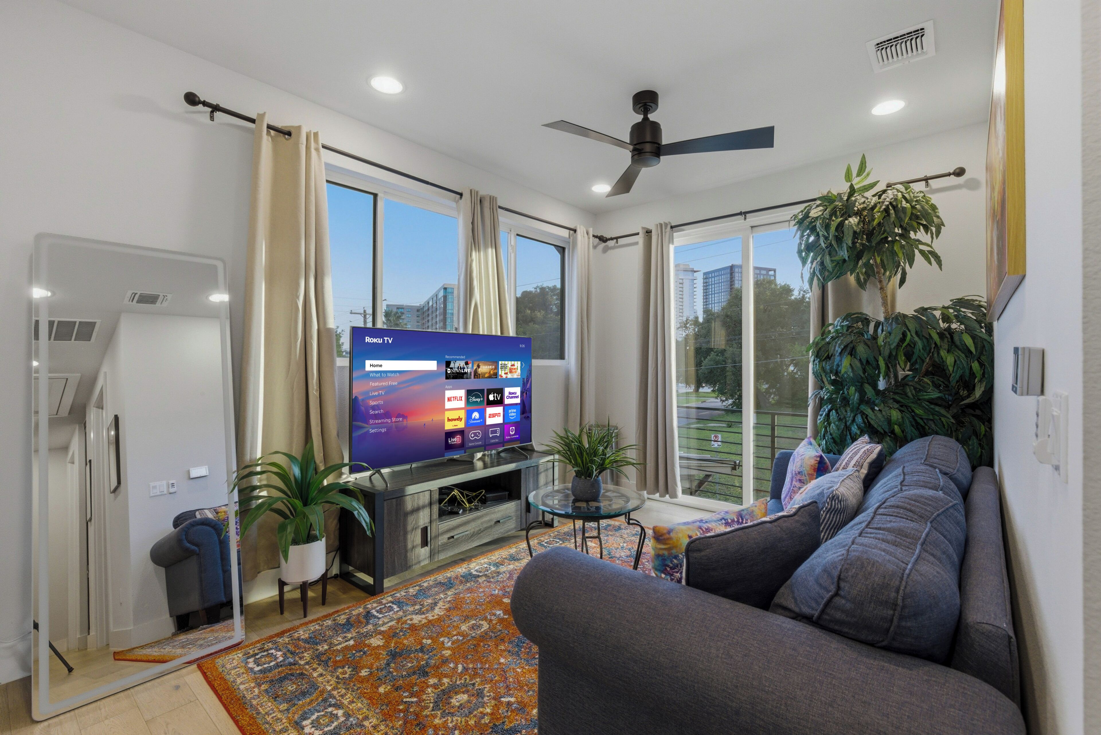 A modern living room with a flat-screen TV, a ceiling fan, a sofa, a coffee table, and a large window.