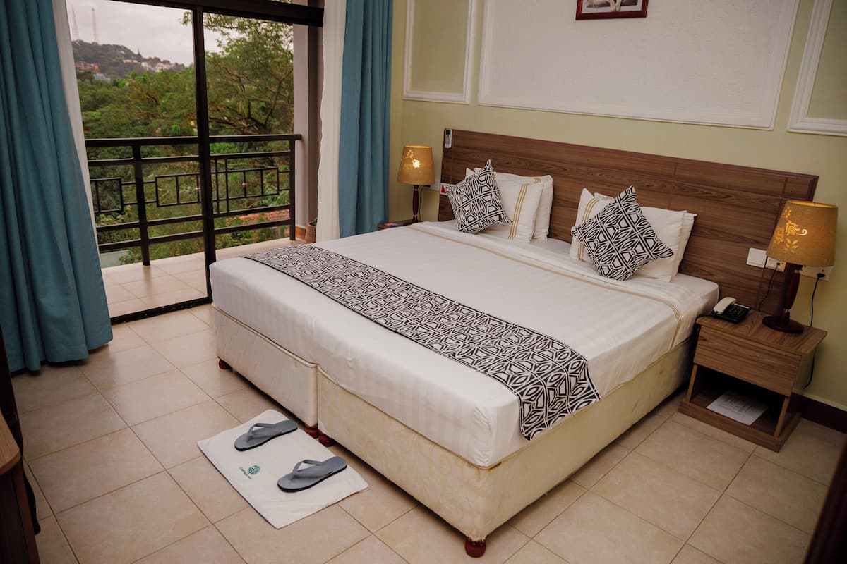 A hotel room with a large bed, two bedside tables with lamps, a view of greenery through a balcony door, and a pair of slippers on a mat.
