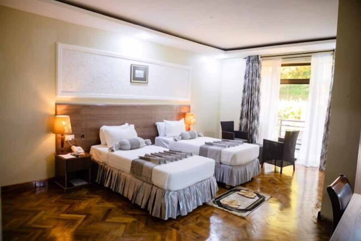 A hotel room with two beds, a desk, a chair, a nightstand with a lamp, and a view of the outdoors.