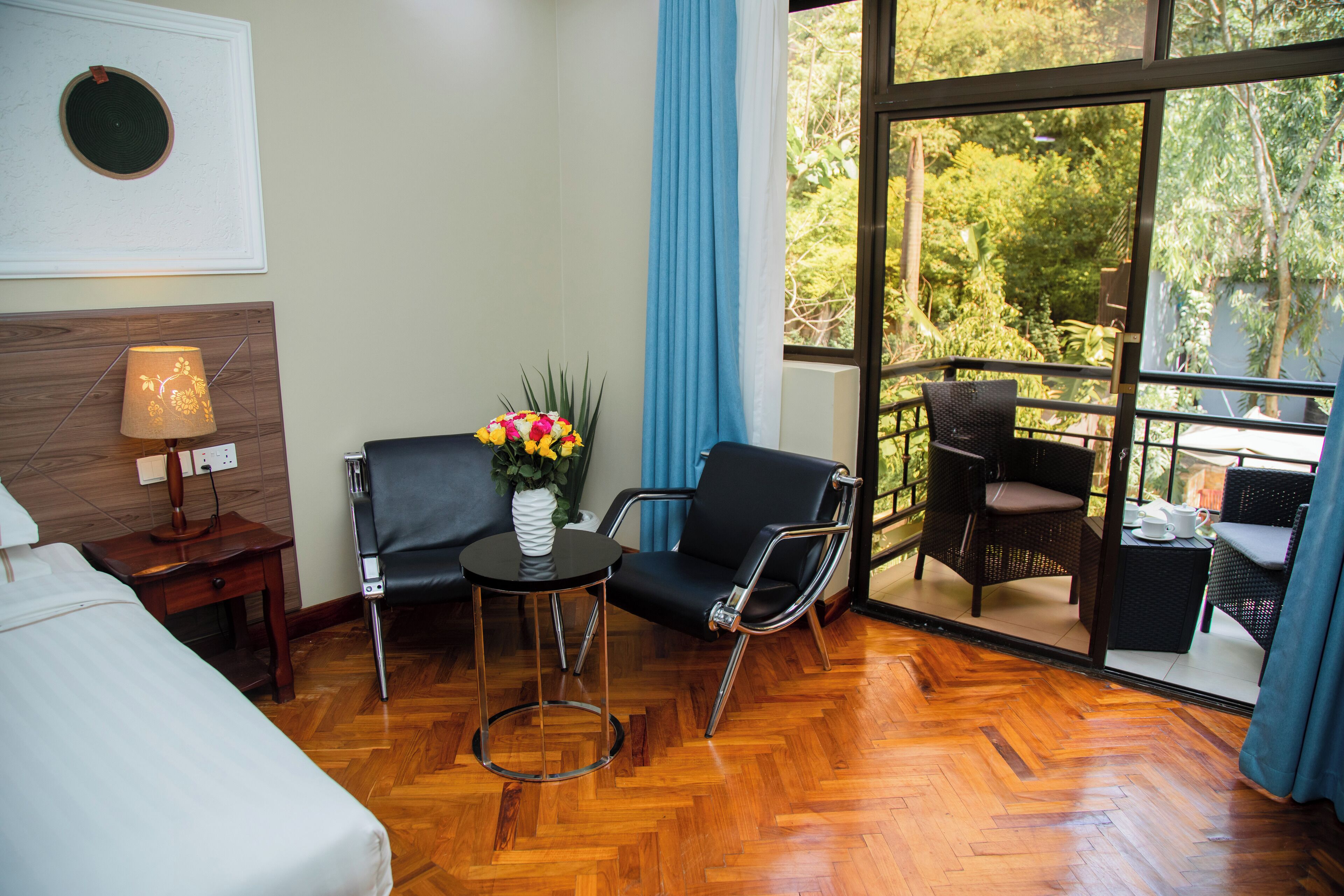A hotel room with a bed, a desk with a lamp, a chair, a small table with a vase of flowers, and a balcony with outdoor furniture.