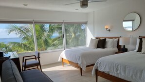 Ein Hotelzimmer mit zwei Betten, einem Schreibtisch, einem Stuhl und einem großen Fenster mit Blick auf Palmen und das Meer.
