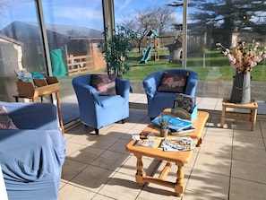 Un patio soleado con dos sillones azules, una mesa de centro de madera y vistas a un jardín trasero con un tobogán y árboles.