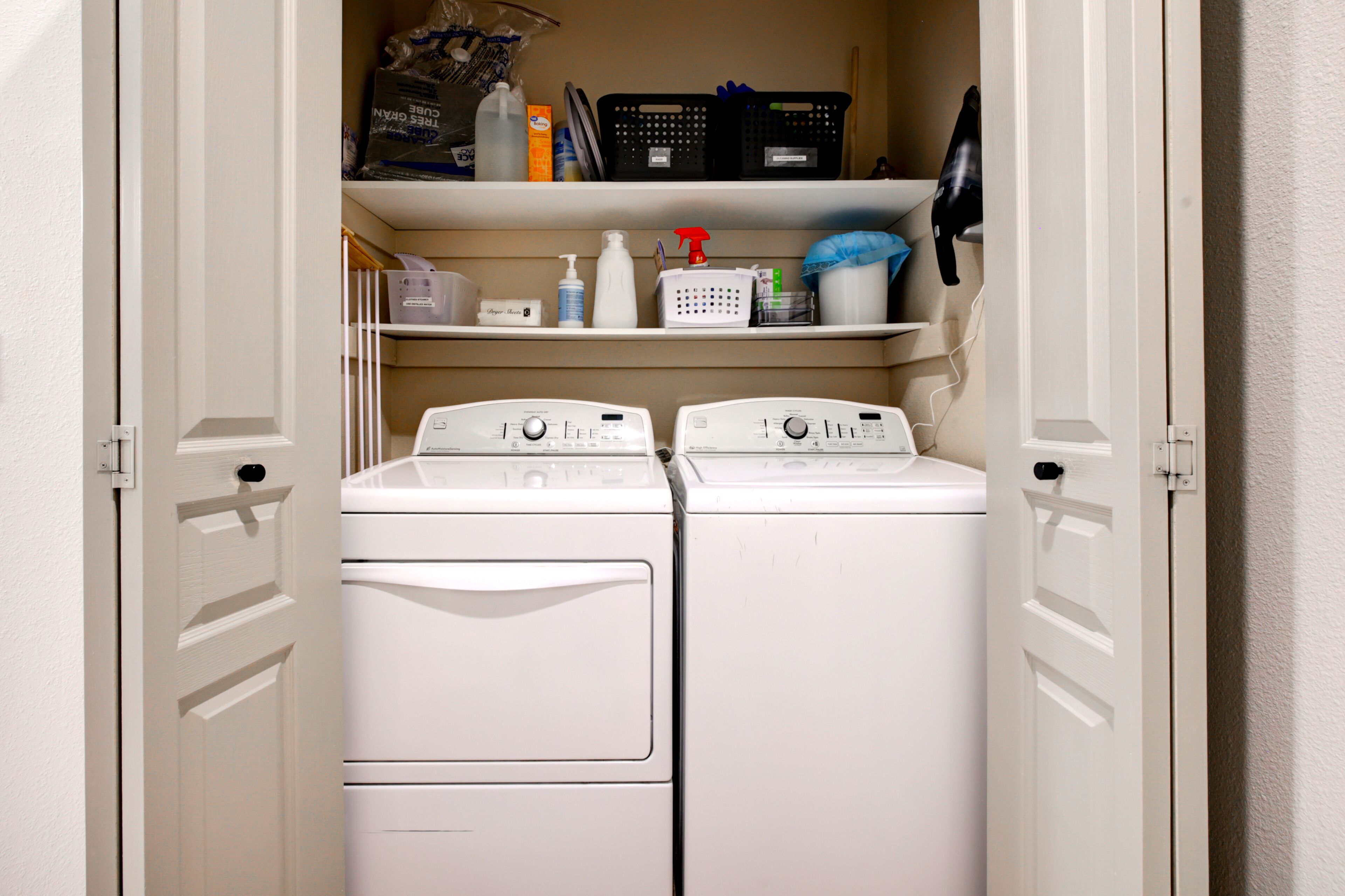 A laundry area with two white front-loading washers and dryers, shelves above containing cleaning supplies and laundry baskets.