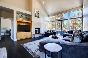 A modern living room with a large TV, comfortable seating, a fireplace, and large windows offering a view of the outdoors.