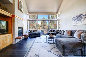A spacious living room with a large TV, comfortable seating, and a view of trees through large windows. - Beautiful Eagle Crest Condo, HOT TUB, 2 Bed/2 Bath (Redmond)