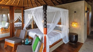 A room with a bed under a canopy, a small table with a lamp, and a view of another room. - Suricata Boma Lodge (Lake Manyara National Park)