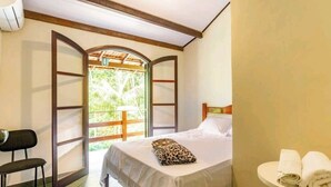 Ein Schlafzimmer mit einem Bett, einem Sessel, einem kleinen Tisch mit Handtüchern und Blick ins Grüne durch eine offene Tür.