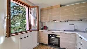 A modern kitchen with wooden cabinets, a built-in oven, a sink, and a window with a view of greenery. - Chez Jacotte (Gérardmer)