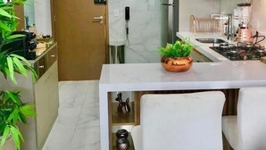 Cozinha moderna com bancada de mármore, fogão embutido e máquina de café. Há dois bancos brancos e um vaso de planta no balcão.