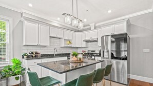 A modern kitchen with white cabinets, a granite countertop, and stainless steel appliances. There are three green bar stools and a central island with a decorative tray.