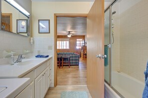 Une salle de bain avec une douche, un lavabo et un miroir. Une porte donne sur un espace de vie comprenant un canapé et une table.