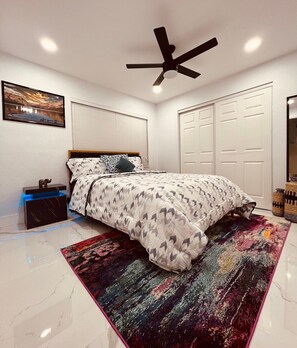 Un dormitorio con cama, mesita de noche, ventilador de techo, ventana grande y puerta.