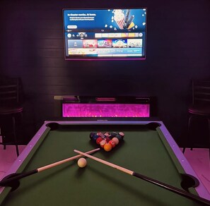 Una mesa de billar con bolas y tacos, un televisor que muestra un canal de cine, y una zona de bar con iluminación de neón.