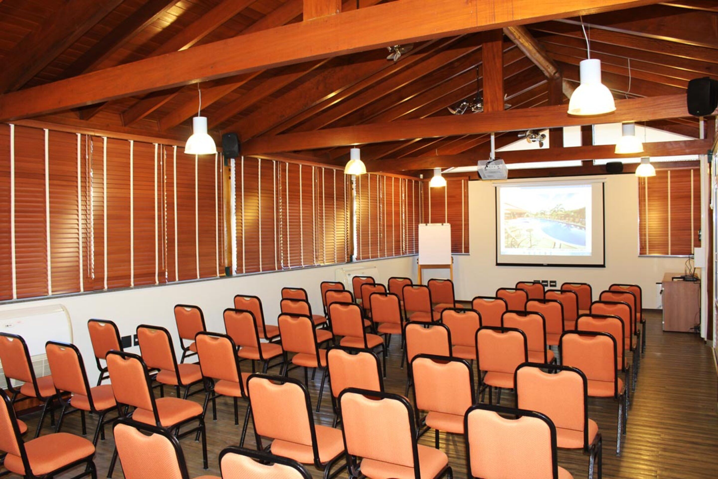 Un salón de conferencias con hileras de sillas color naranja, vigas de madera en el techo y una pantalla de proyector en la pared.