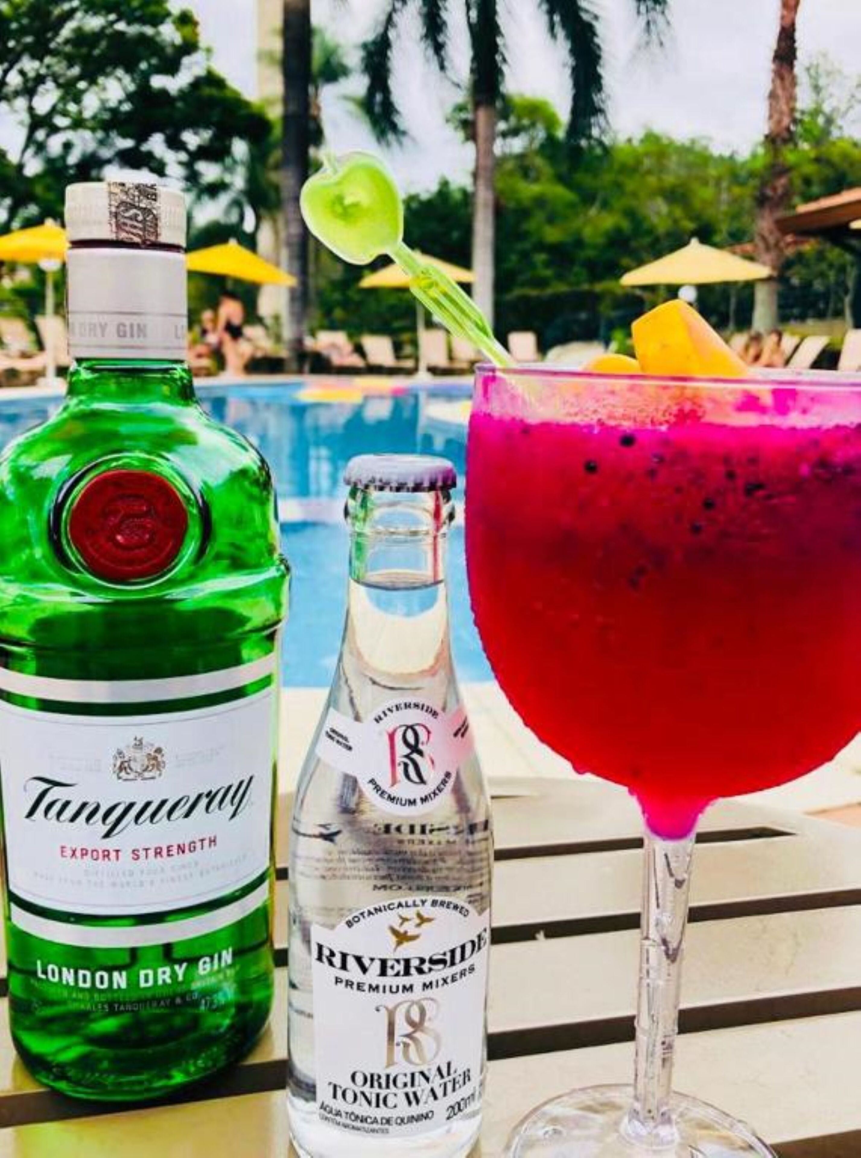 A bottle of Tanguey London Dry Gin and a glass of pink cocktail with a lime garnish, set against a poolside backdrop.