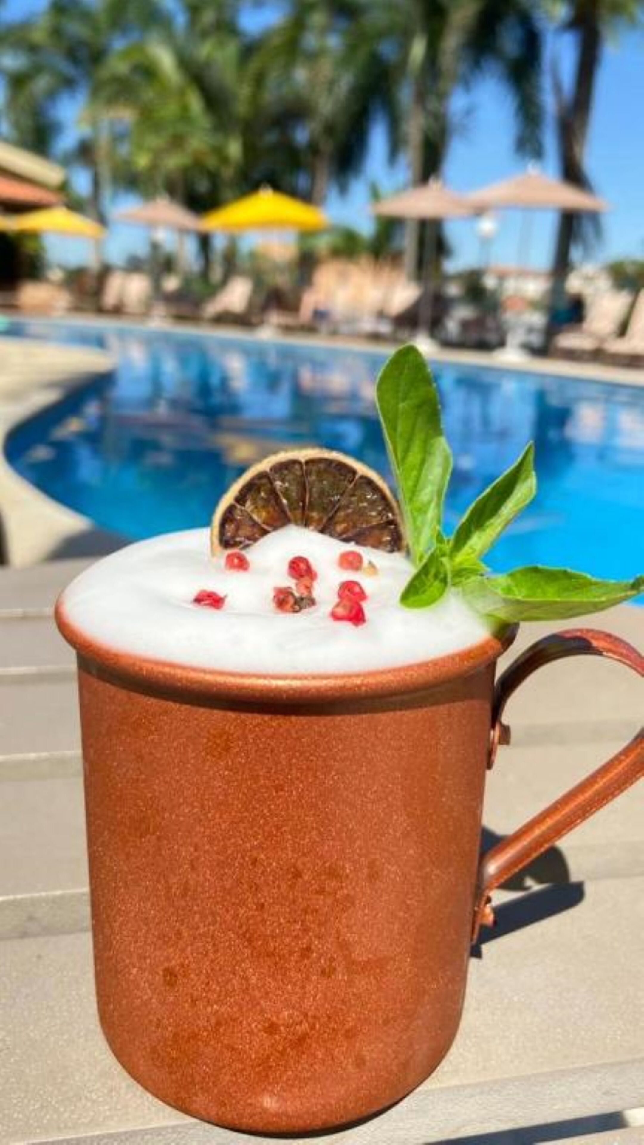 A mug with a creamy beverage, garnished with a slice of fruit and a leaf, placed on a poolside table with a pool and umbrellas in the background.