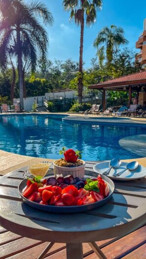 A poolside table with a fruit bowl and a dessert, surrounded by palm trees and a pool. - LS Villas Hotel & SPA (Aguas de Sao Pedro)