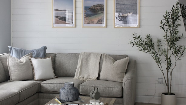 Ein Wohnzimmer mit grauem Ecksofa, drei gerahmten Bildern an der Wand, einer Topfpflanze und einem Couchtisch mit Dekoration.