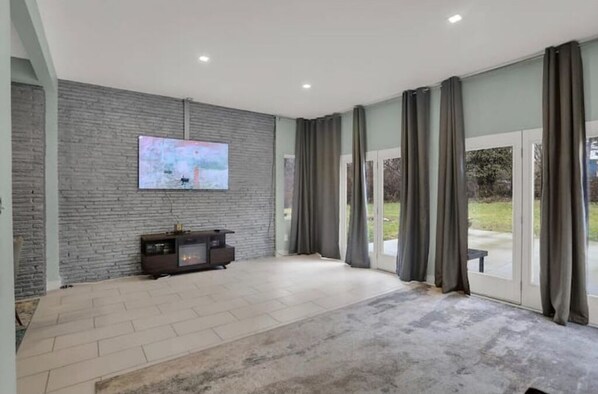 A spacious living room with a flat-screen TV mounted on a brick wall, a dark entertainment unit, and a large sliding glass door leading to an outdoor area. - MidCentury Modern Mansion (Indianapolis)