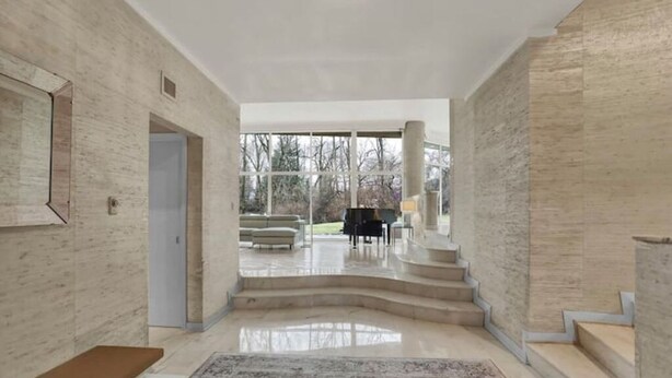 Un intérieur spacieux avec un magnifique escalier menant à un balcon, un piano et un coin salon offrant une vue sur l’extérieur.