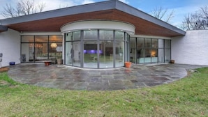 Une maison moderne dotée d’une entrée en verre incurvé, d’une terrasse en pierre et d’un toit en bois.