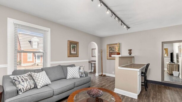 A living room with a grey sofa, a wooden coffee table, and a kitchen area with a counter and sink. - The Lewis & Clark Hangout (Saint Charles)
